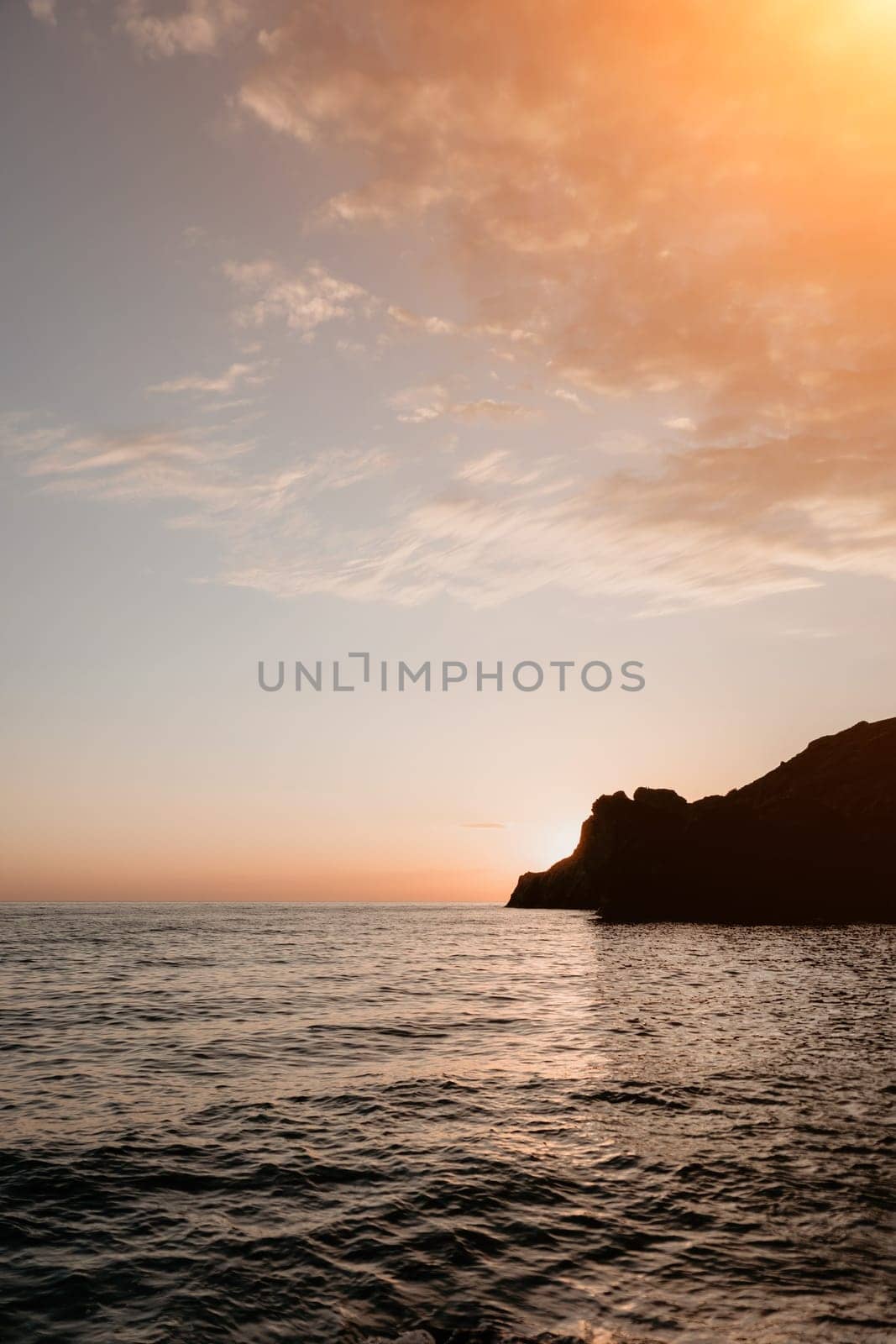 A red burning sunset with the silhouette of a cliff over the sea. Abstract nature summer or spring ocean sea background. Small waves on golden warm water surface with bokeh lights from sun.