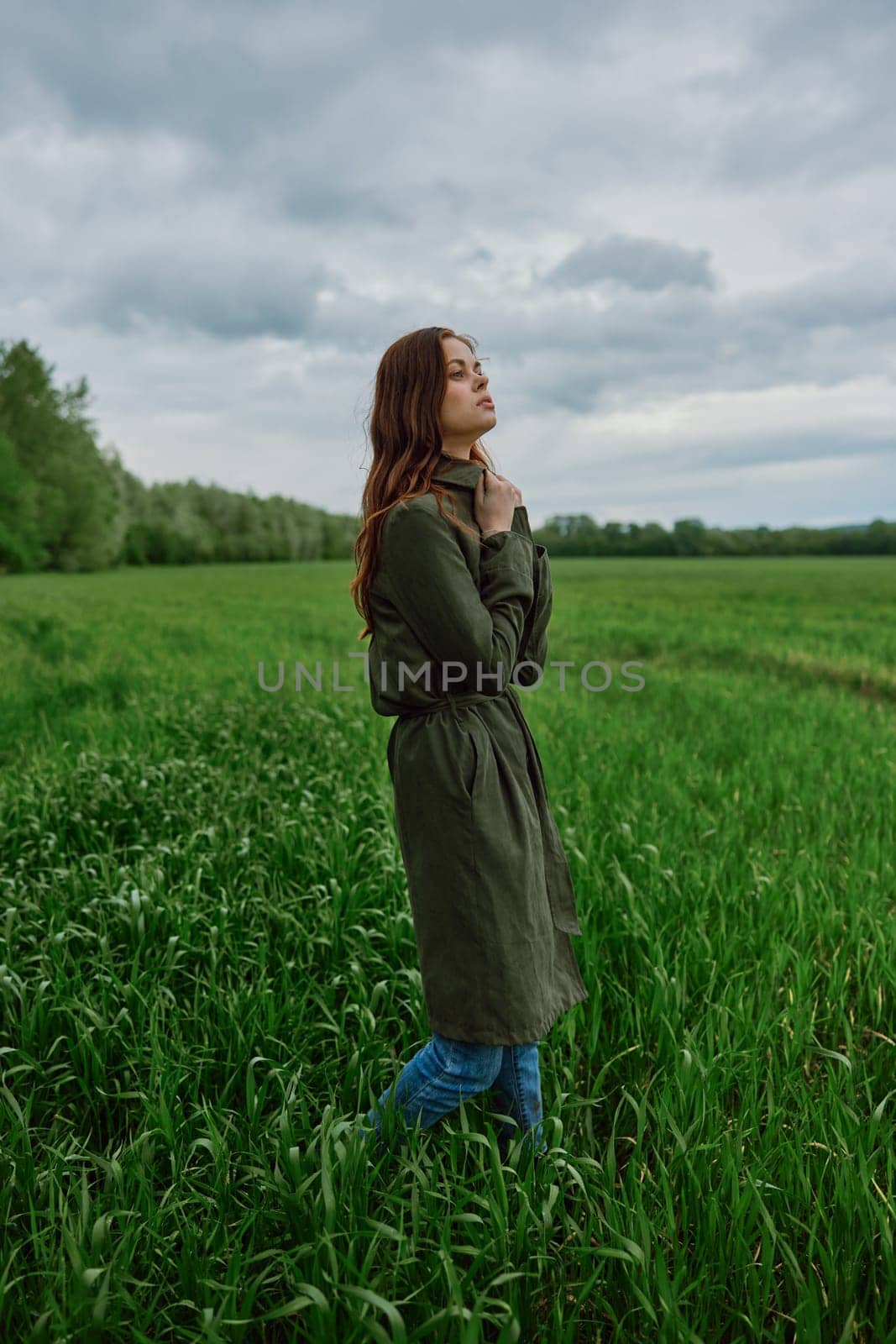 a beautiful woman in a dark coat stands in a green field in the spring in rainy weather. High quality photo