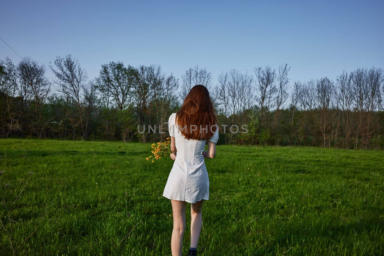 a woman with long red hair in a white dress stands in a green field at sunset with her back to the camera by Vichizh