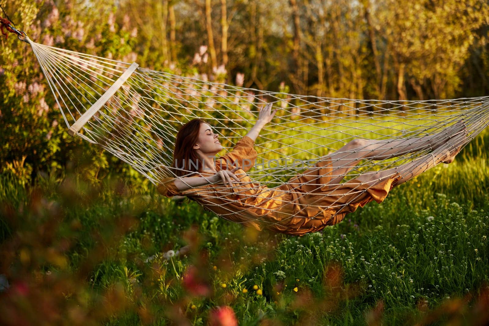 a beautiful woman is resting in nature lying in a mesh hammock in a long orange dress looking to the side, arms outstretched. Horizontal photo on the theme of recreation. High quality photo