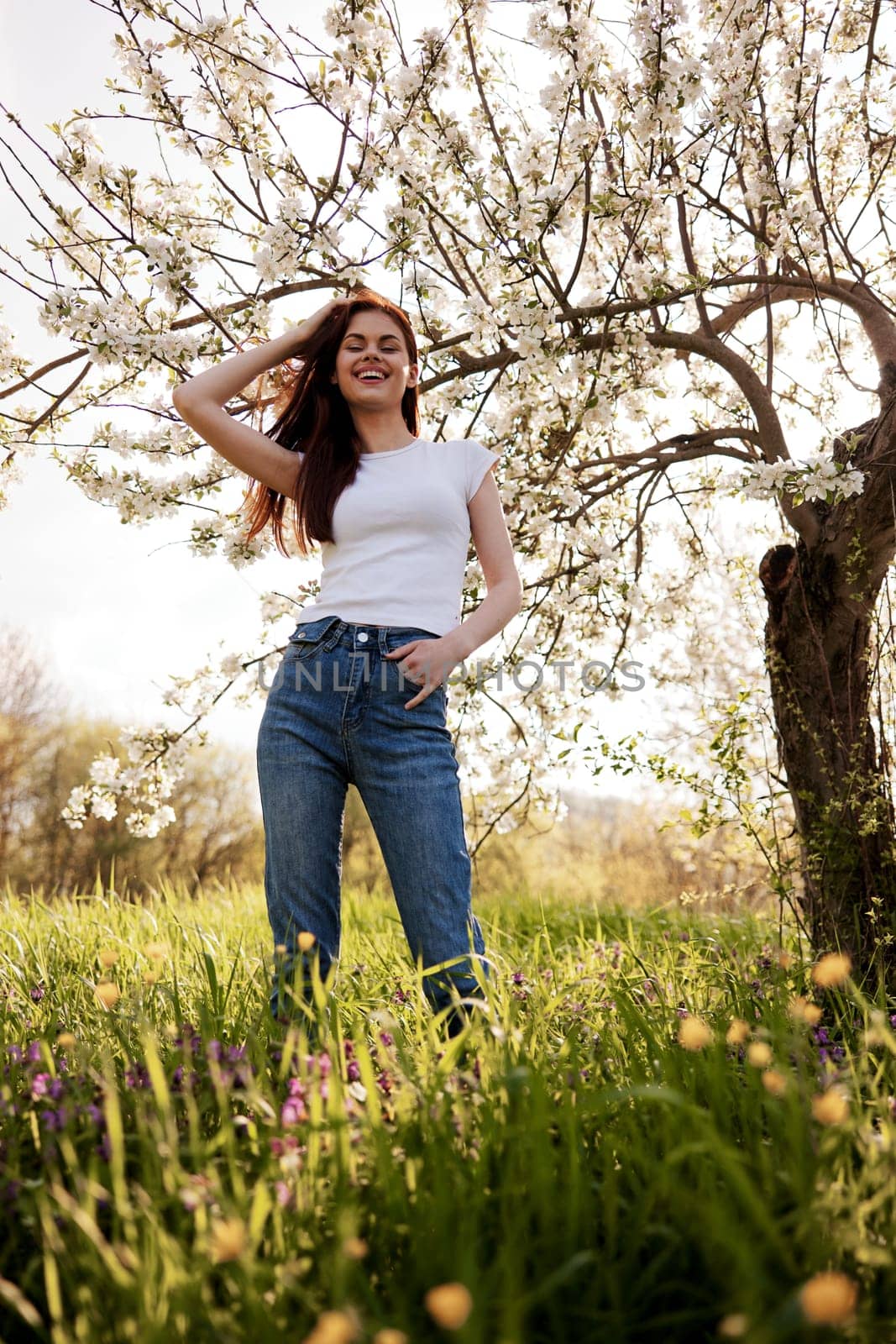 joyful, happy woman in jeans and a light T-shirt posing against the backdrop of a flowering tree by Vichizh