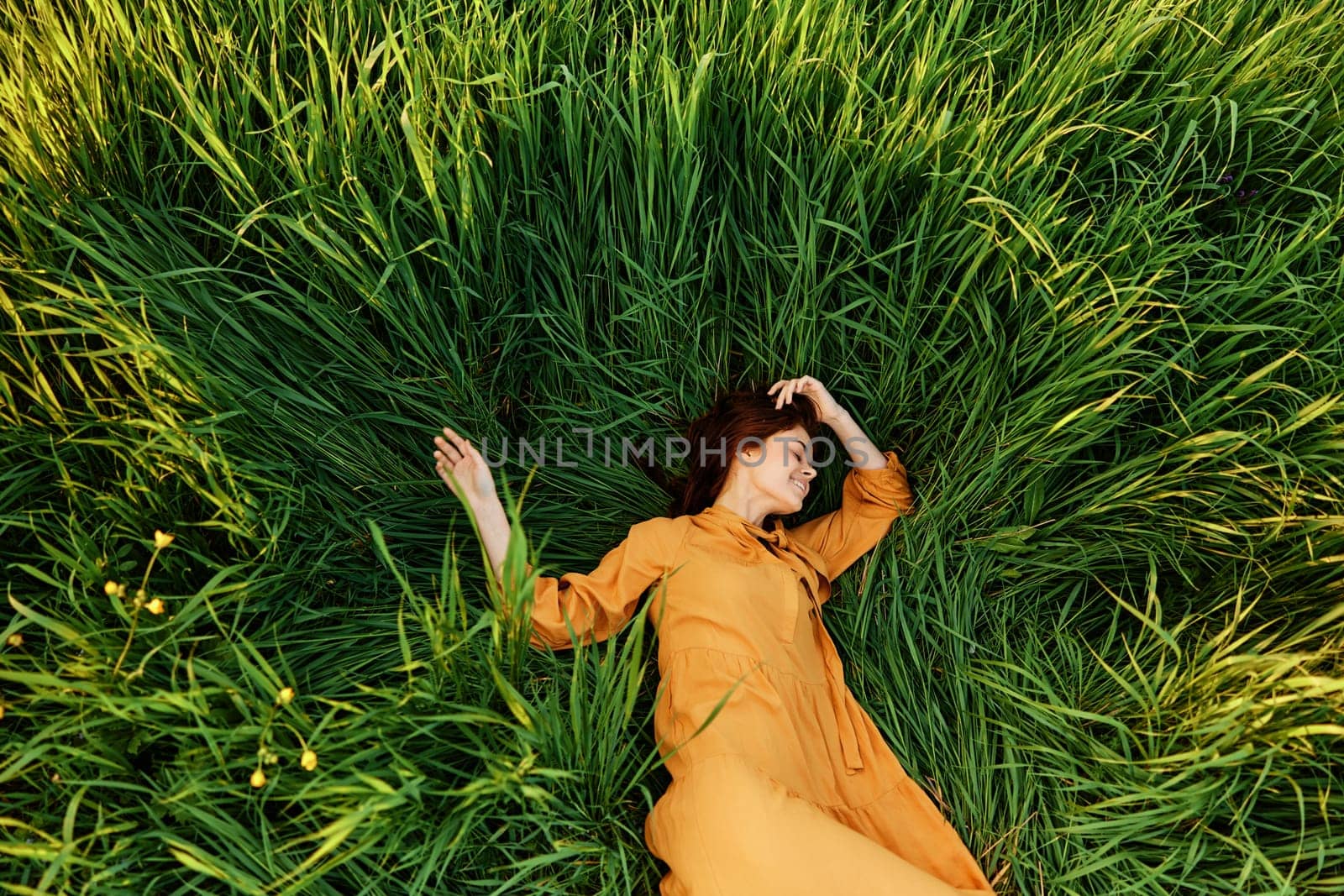 an elegant woman lies in the tall green grass in a long summer orange dress and with her arms outstretched enjoys nature and sunny weather with her eyes closed by Vichizh