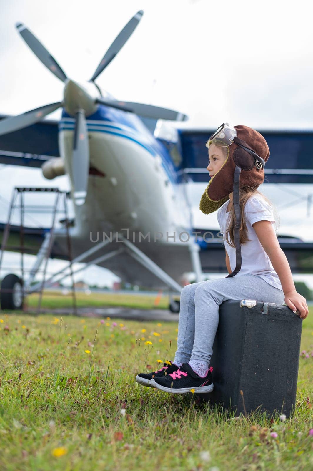 A little girl in a pilot's costume sits on a suitcase against the background of an airplane with a propeller. Child Dreams of flying in the sky by mrwed54