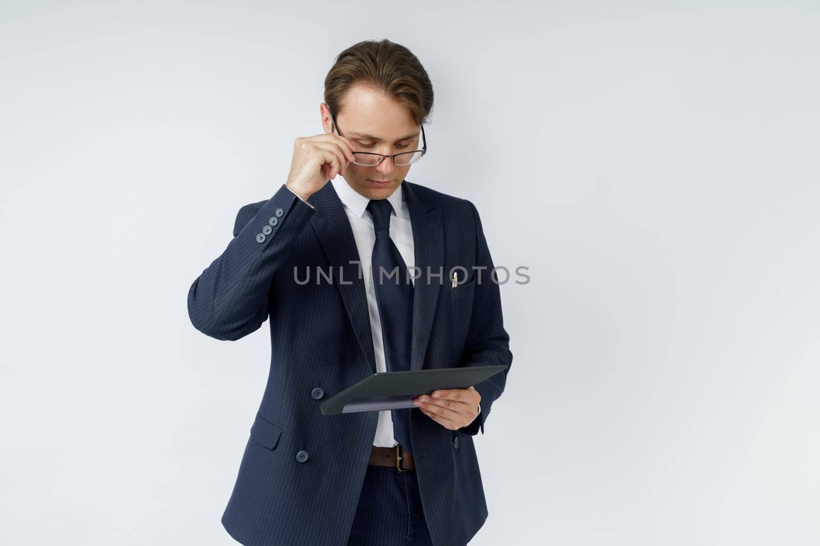 Portrait of a businessman in a blue suit who is holding an electronic tablet in his hands and adjusting his glasses. White background. Business and finance concept