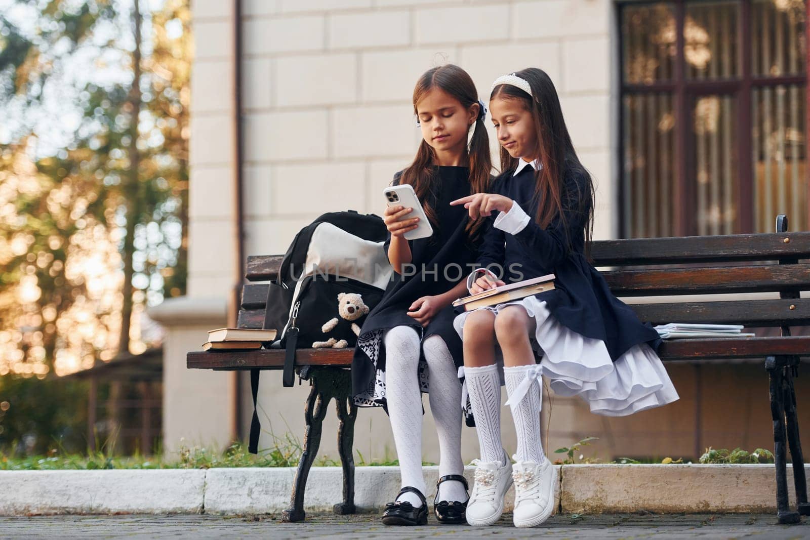 Two schoolgirls is sitting outside together near school building.