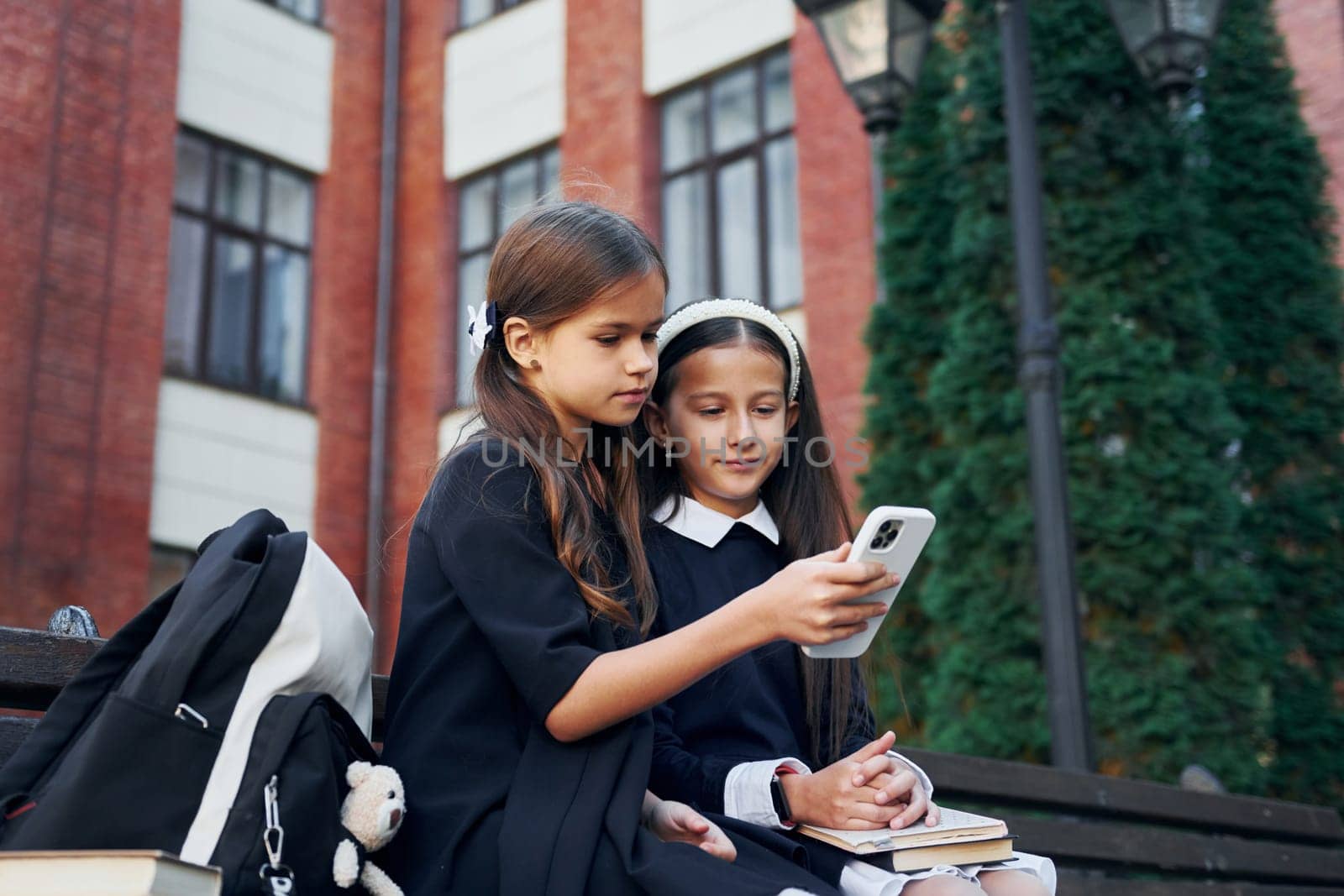 Sitting together. Two schoolgirls is outside near school building.