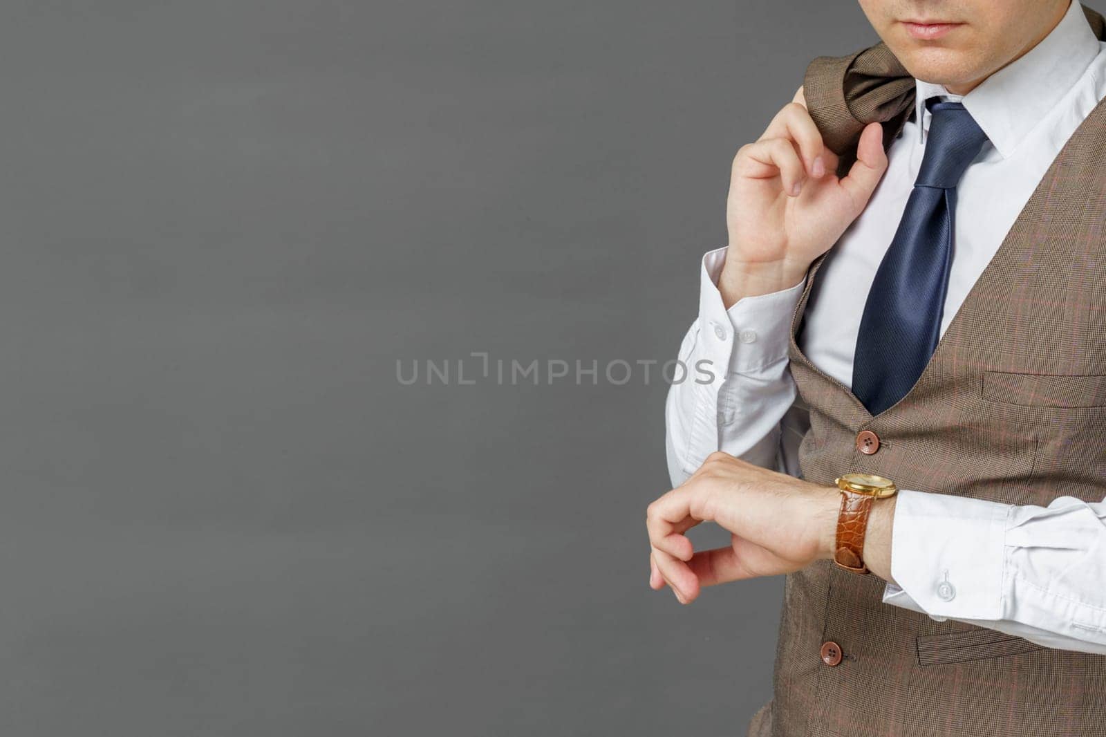 Portrait of a businessman who looks at his watch and holds a jacket over his shoulder. The face is not visible. Gray background. Business and finance concept