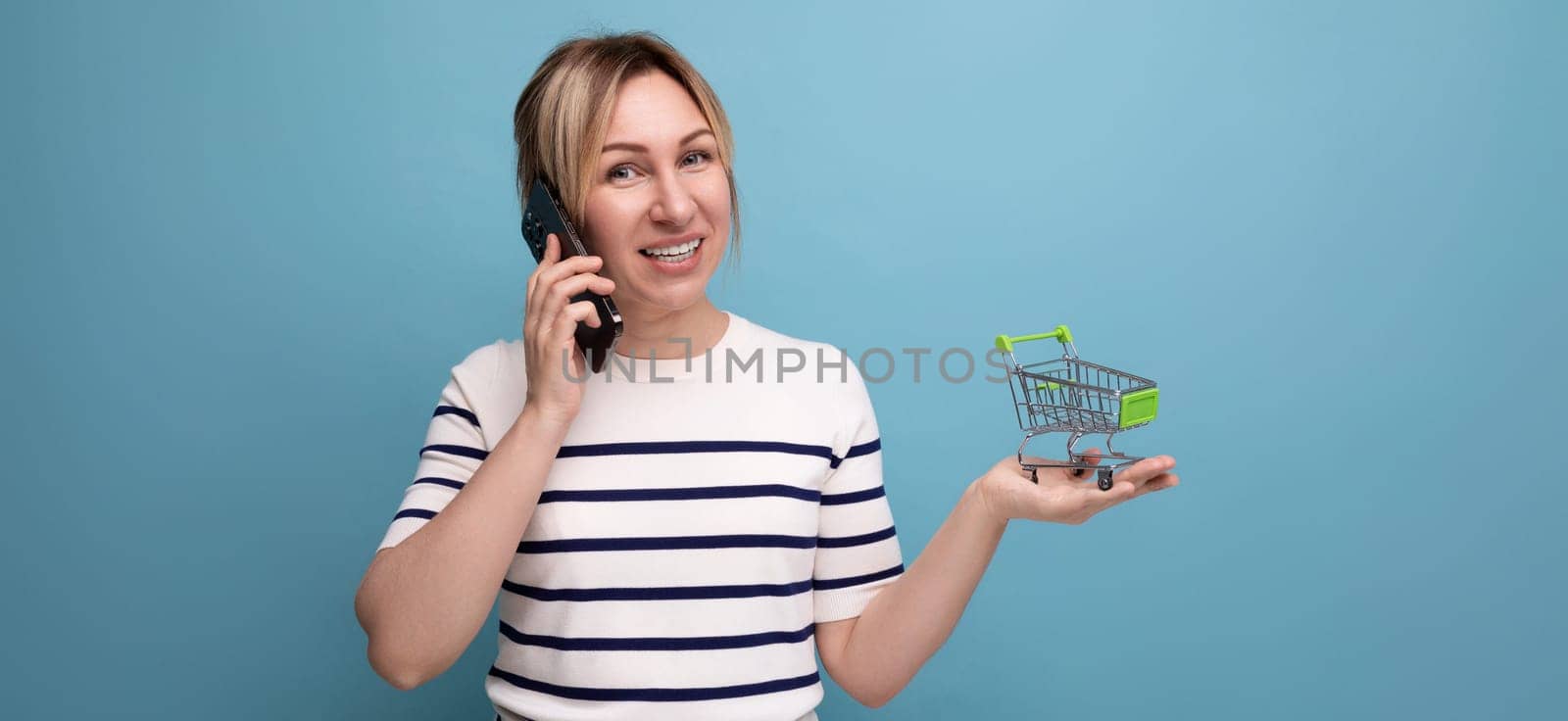 panoramic photo of young cute casual woman shopaholic talking on phone holding empty shopping cart in supermarket on blue background with copy space.