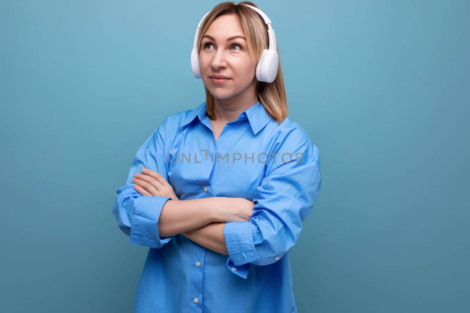 charming european girl in a casual shirt listens to music in big white headphones on a blue isolated background by TRMK