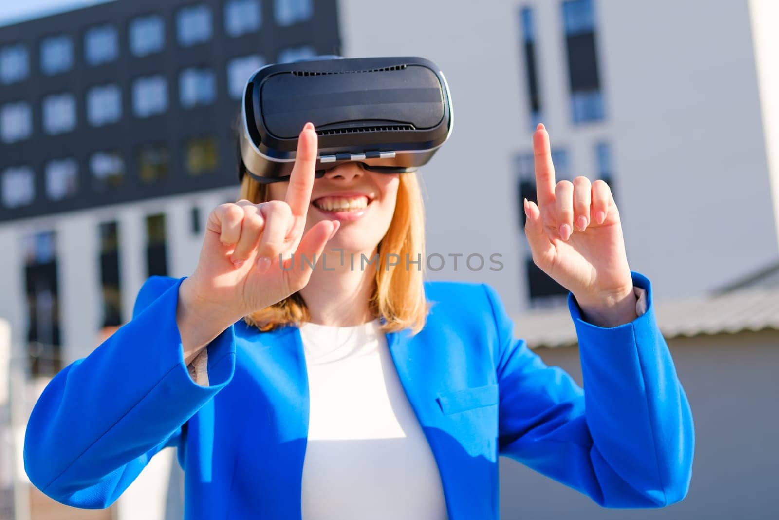 A happy woman in the suit is standing outside and gestures in augmented reality in 3D VR goggles.