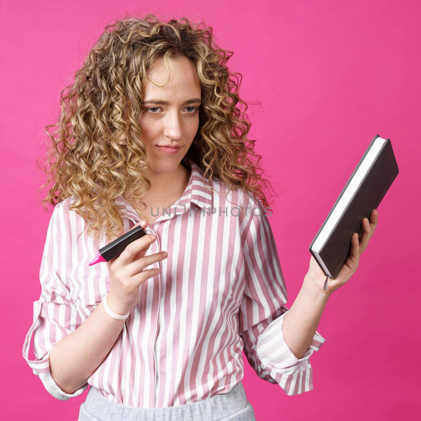 Fashionable woman in a striped shirt holding a diary and a marker. Isolated on pink background