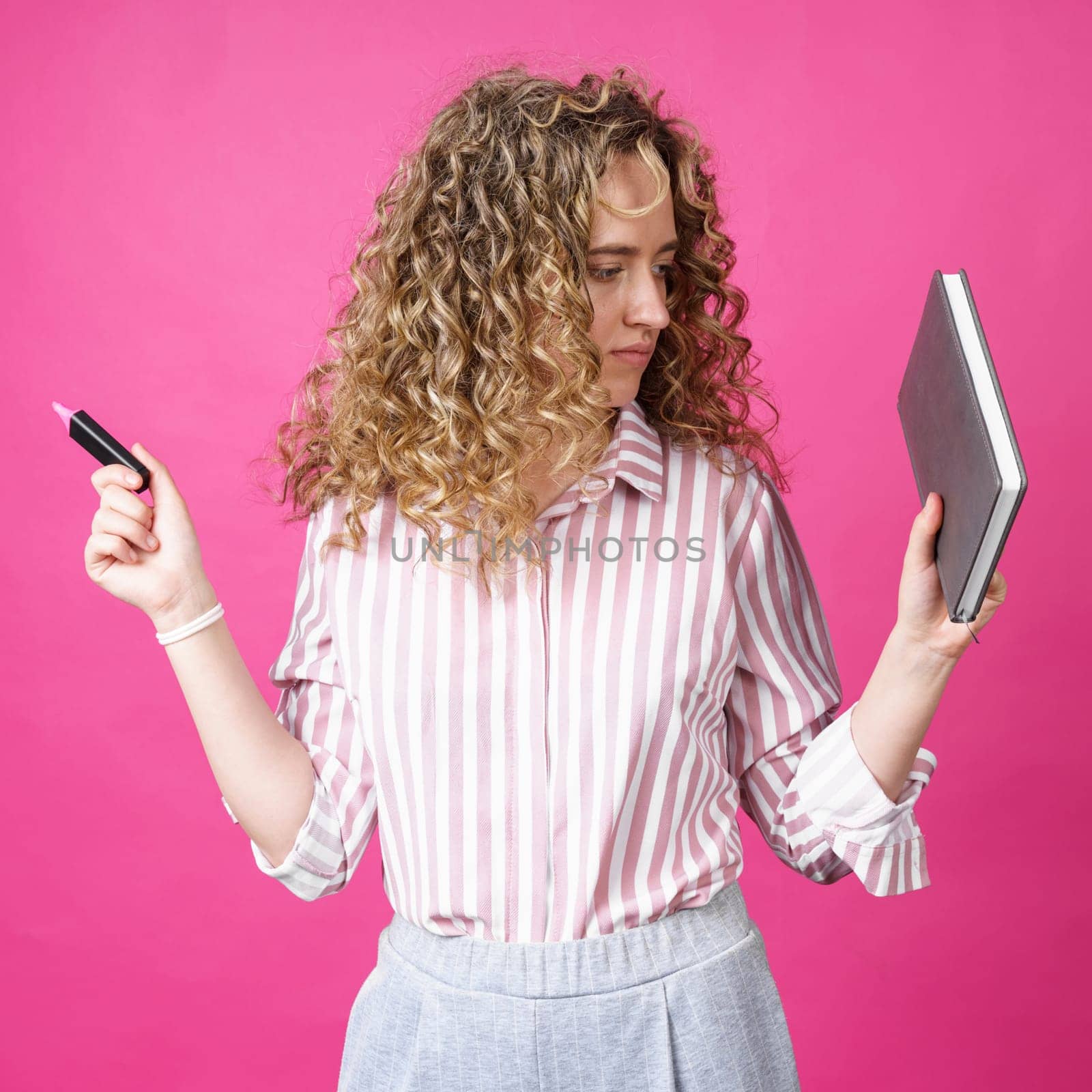 Fashionable woman in a striped shirt holding a diary and a marker. Isolated on pink background