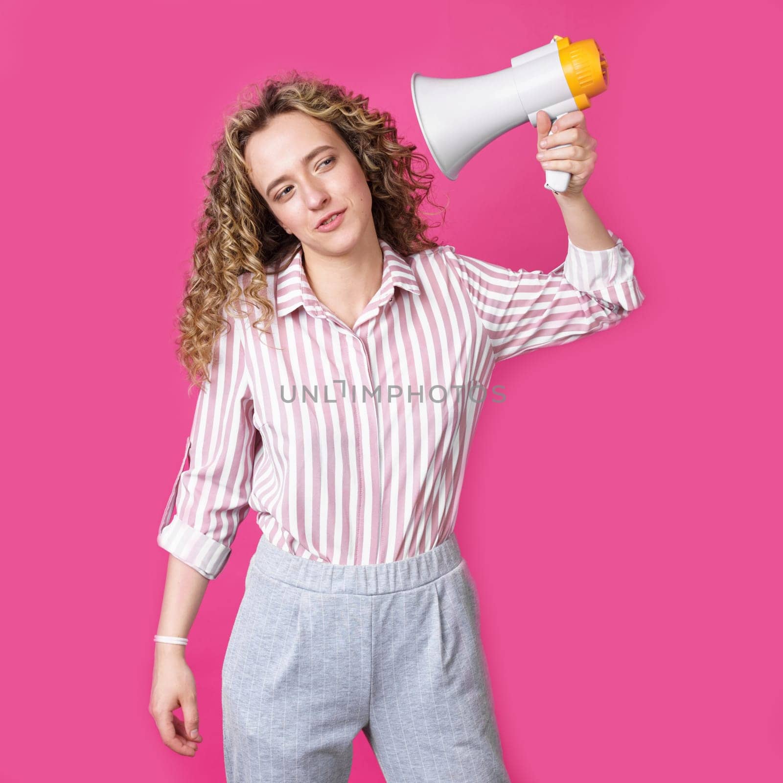 A young woman holds a screaming megaphone in her hands, which is aimed at her face. Isolated pink background. People sincere emotions lifestyle concept.