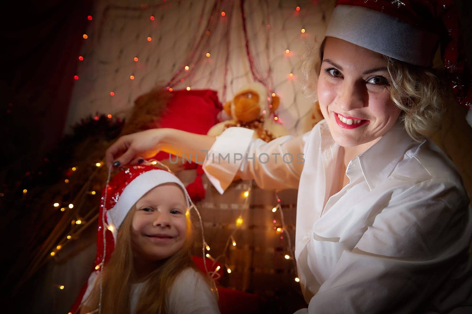 Cute mother and daughter in the red hats of Santa Claus assistants in a room decorated for Christmas. The tradition of decorating house and dressing up for holidays. Happy childhood and motherhood
