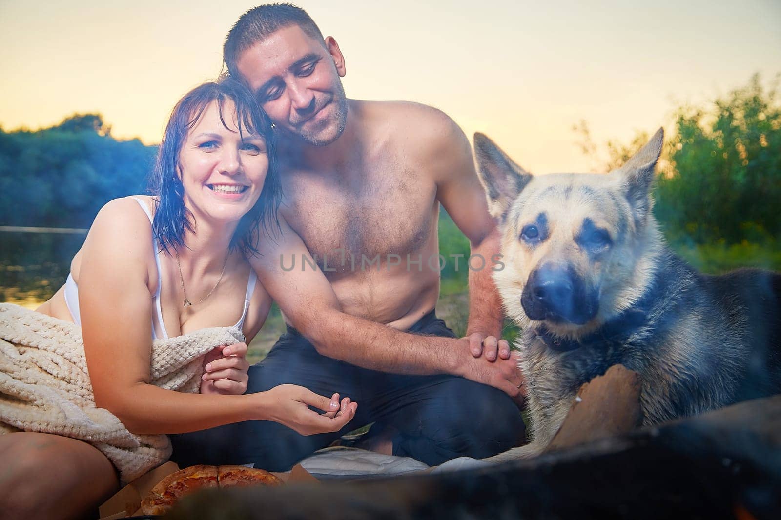 Happy wet couple relaxin, having fun and hugs with big dog near fire in camping on nature at summer sunny evening in sunset. Family or lovers have date and rest outdoor. Concept of love by keleny