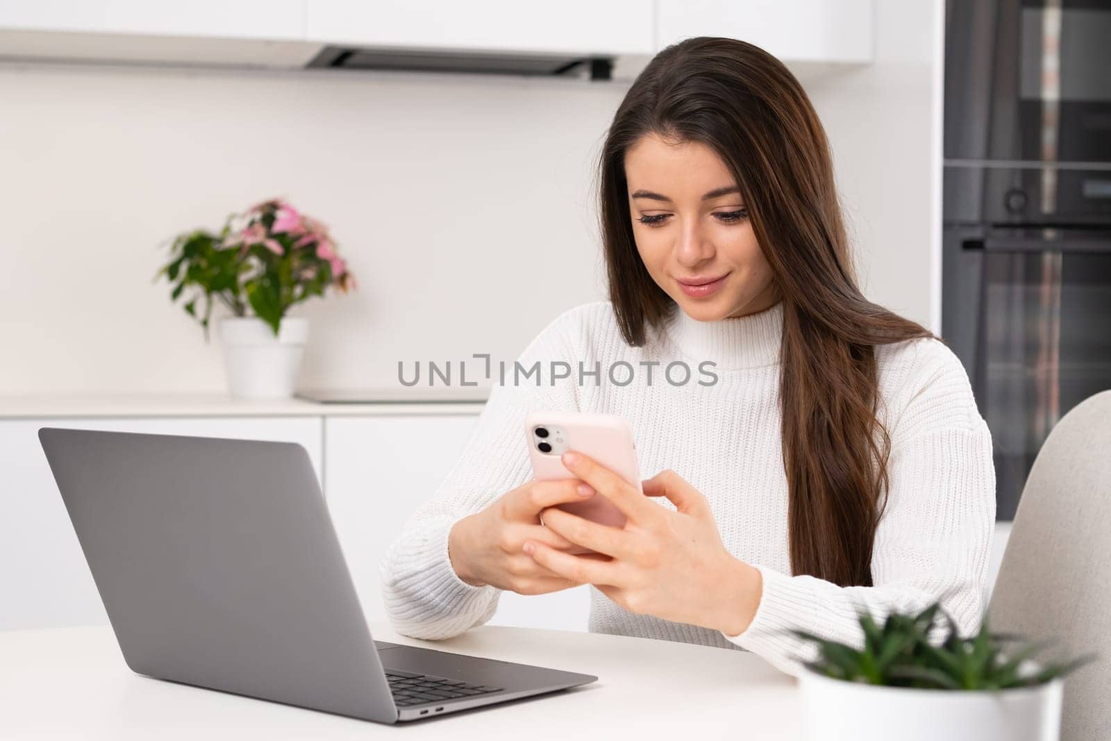 Focused woman types message to boyfriend on smartphone. Brunette lady sitting near notebook and uses her mobile phone at break