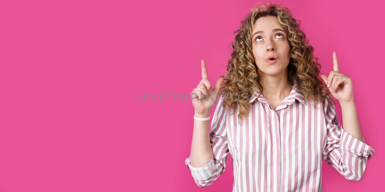 Young beautiful woman, amazed and surprised, looking up and pointing with fingers and raised hands. Isolated on pink background.