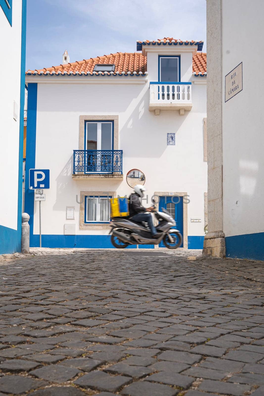 Food delivery moto scooter driver with yellow backpack behind back to deliver food in Ericeira streets , Portugal. by papatonic