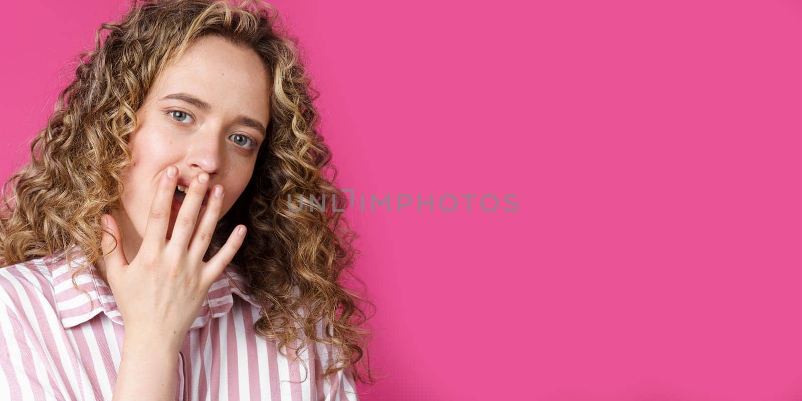 Young beautiful woman covers her face with her hand. Surprised look. Isolated pink background