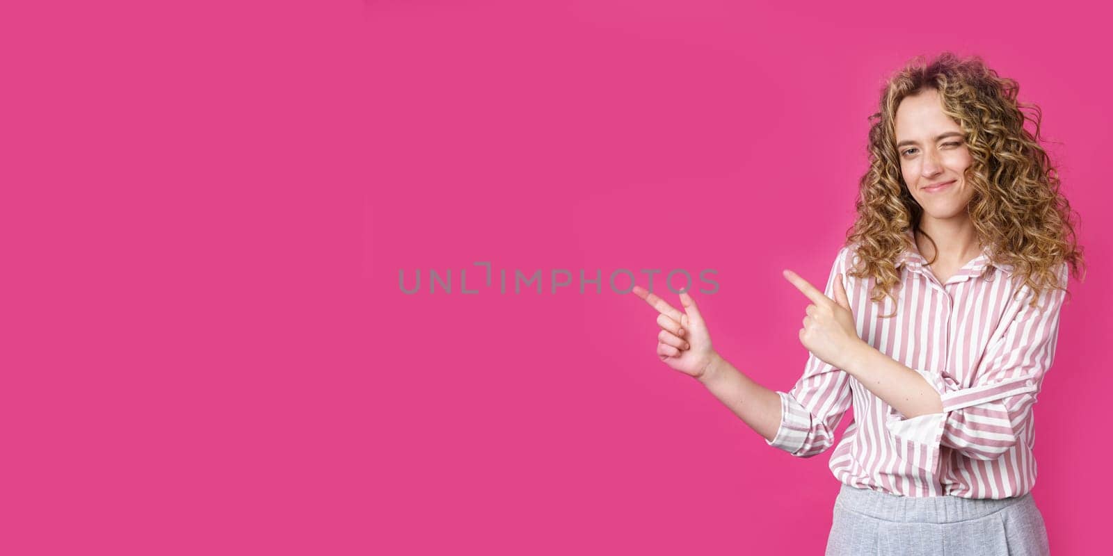 A cheerful woman points to a copy space, discusses an amazing ad, gives way or a direction, wears a striped shirt, has a pleasant smile, and feels optimistic. Isolated on pink background