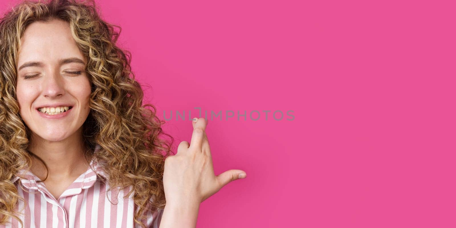 Waiting for a special moment. A portrait of a smiling woman with closed eyes who keeps her fingers crossed. Make a wish. Isolated on a pink background. People sincere emotions concept.