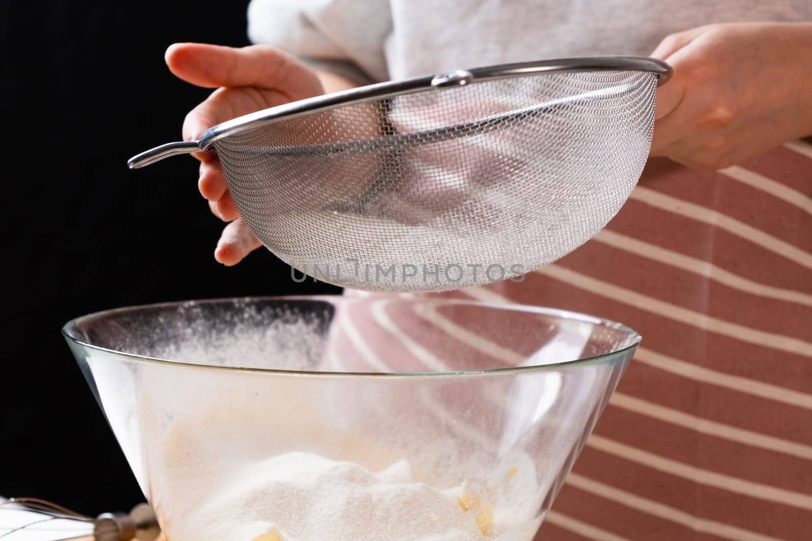 Close up woman sifts wheat flour through a sieve into mixing bowl for dough preparation for cake and bakery on the black background. Knead the dough for homemade pastry.
