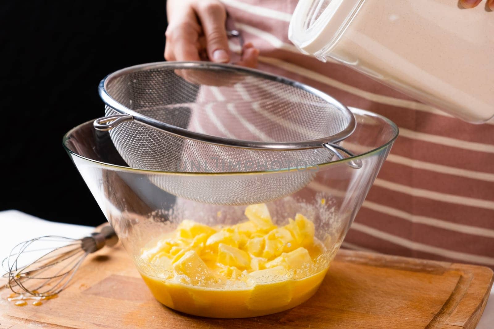 Close up woman sifts wheat flour through a sieve into mixing bowl for dough preparation for cake and bakery on the black background. Knead the dough for homemade pastry.