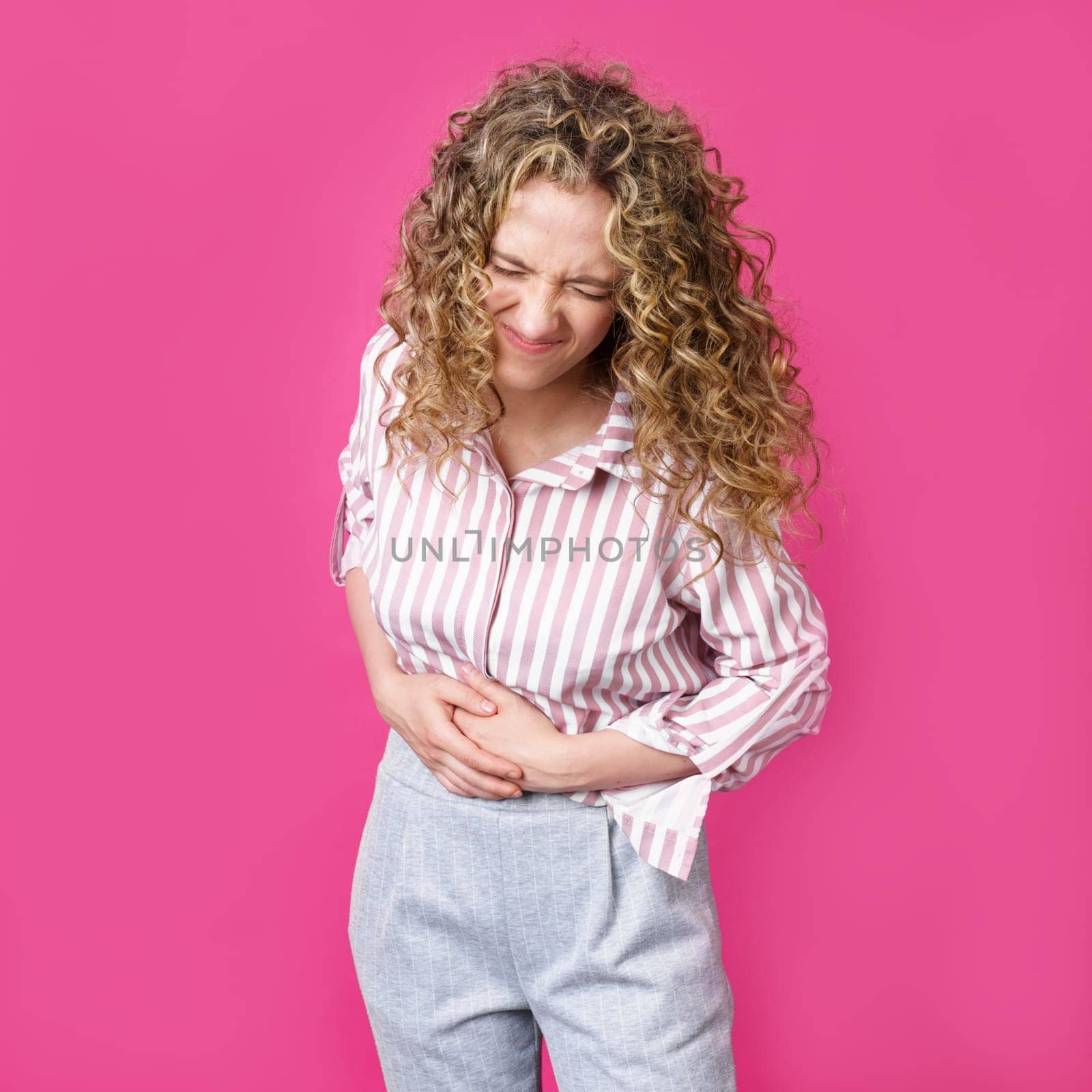 The young woman closed her eyes, laughing holding her belly. Isolated on pink background