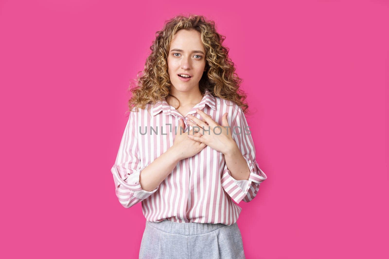 The woman holds her hands on her heart, wearing a striped shirt, expresses gratitude, has a friendly expression on her face. Isolated on pink background