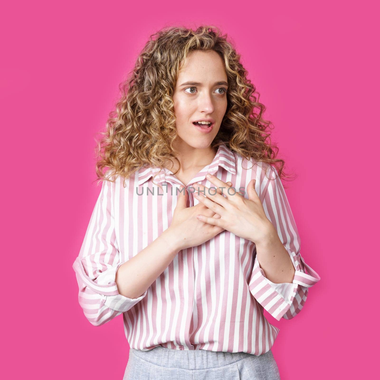 The woman holds her hands on her heart, wearing a striped shirt, expresses gratitude, has a friendly expression on her face. Isolated on pink background