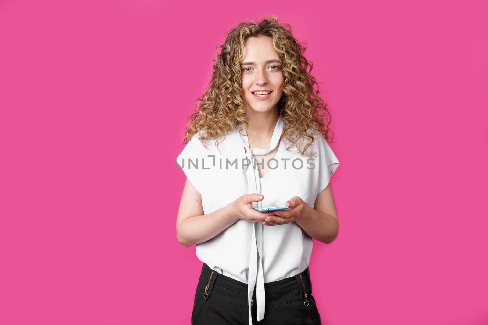 A young woman holds a mobile phone in her hands and smiles. Isolated pink background