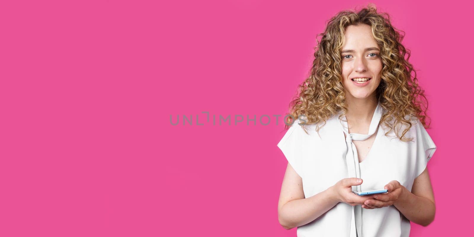 A young woman holds a mobile phone in her hands and smiles. Isolated pink background