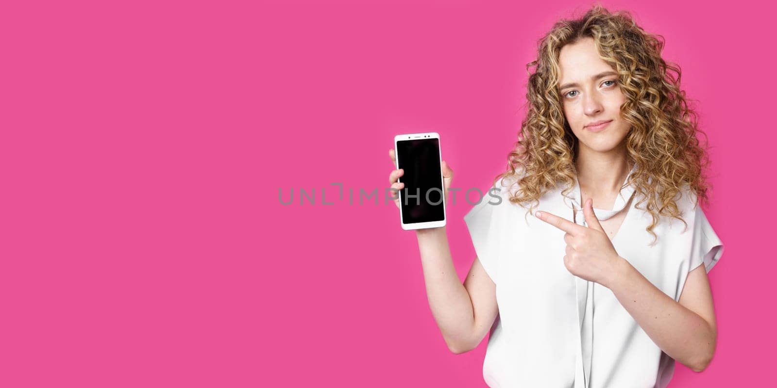 Look at this cell phone. Contented happy woman, pointing her index finger at a blank screen, shows a modern device. Isolated on a pink background. Technology concept