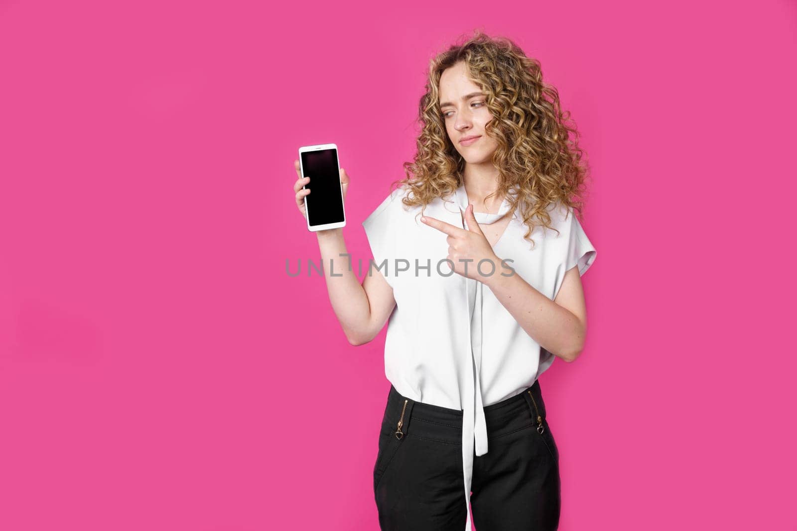 Look at this cell phone. Contented happy woman, pointing her index finger at a blank screen, shows a modern device. Isolated on a pink background. Technology concept