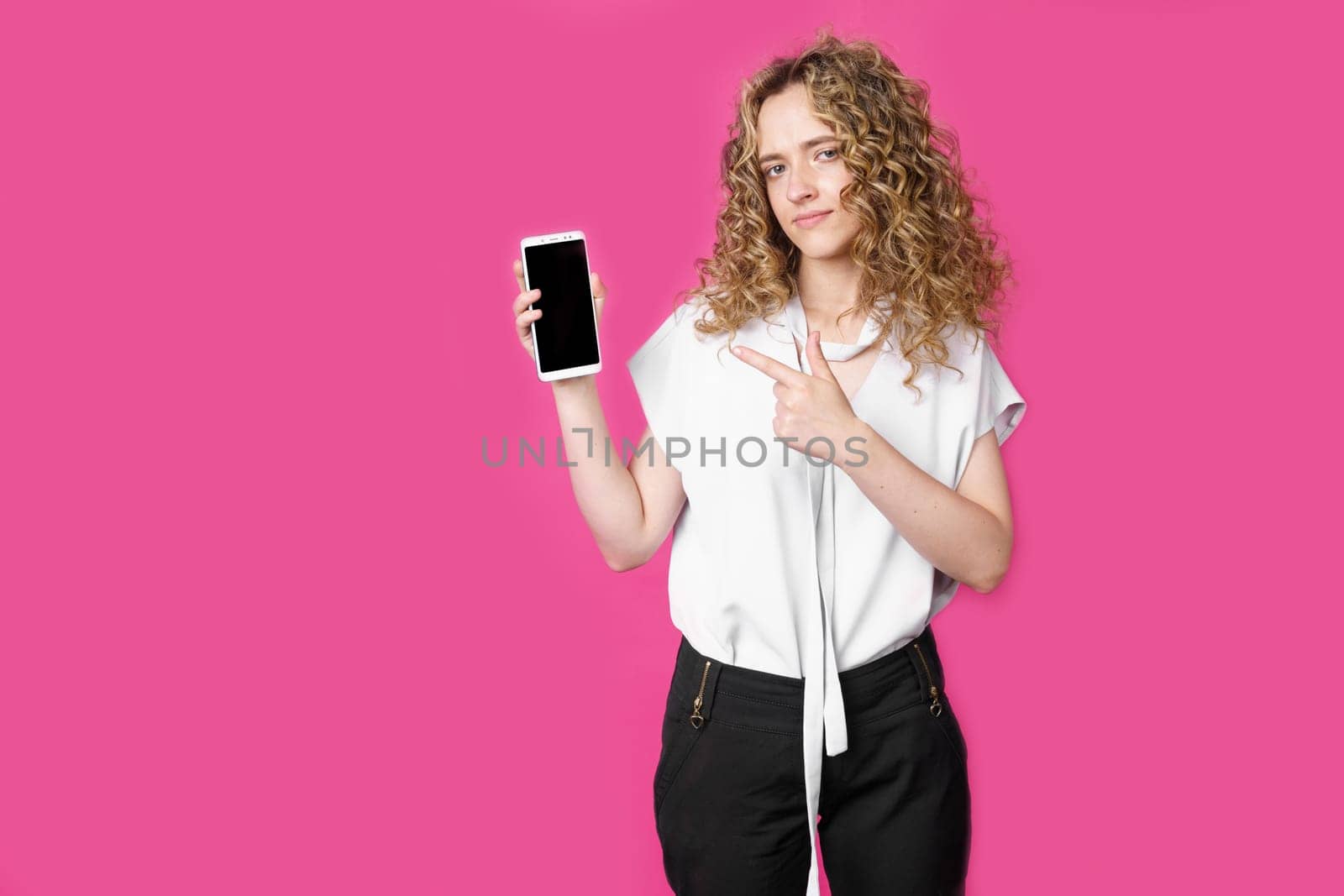 Look at this cell phone. Contented happy woman, pointing her index finger at a blank screen, shows a modern device. Isolated on a pink background. Technology concept