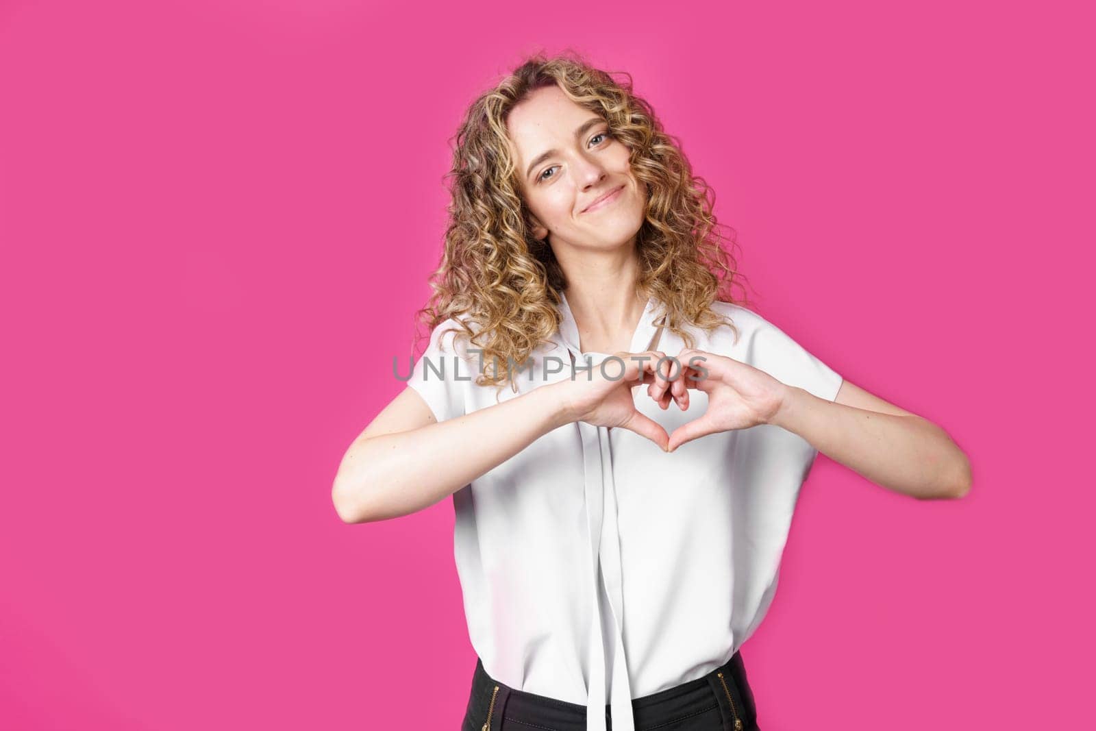 Portrait of a beautiful woman who shows a heart gesture on her chest, passionately, expresses love for a loved one. Isolated pink background.