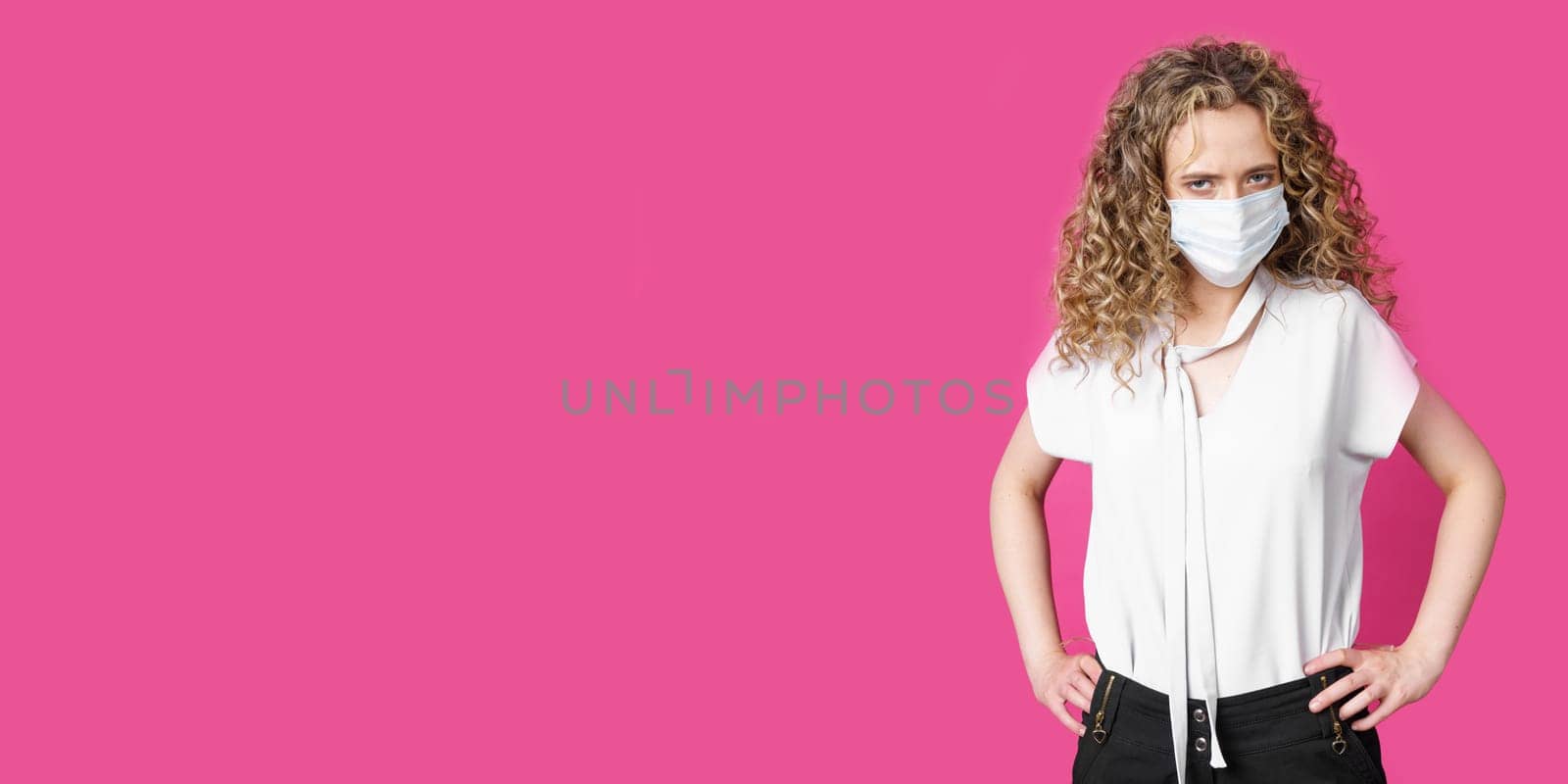 A young woman with a medical mask on her face is standing with her hands on her waist. Isolated pink background