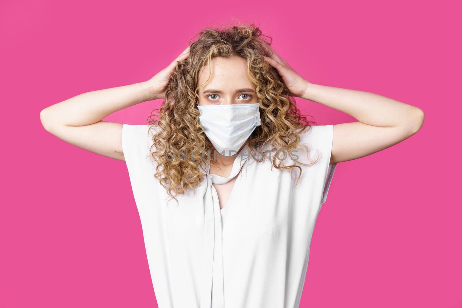 A young woman with a medical mask on her face holds her head with her hands. Isolated pink background