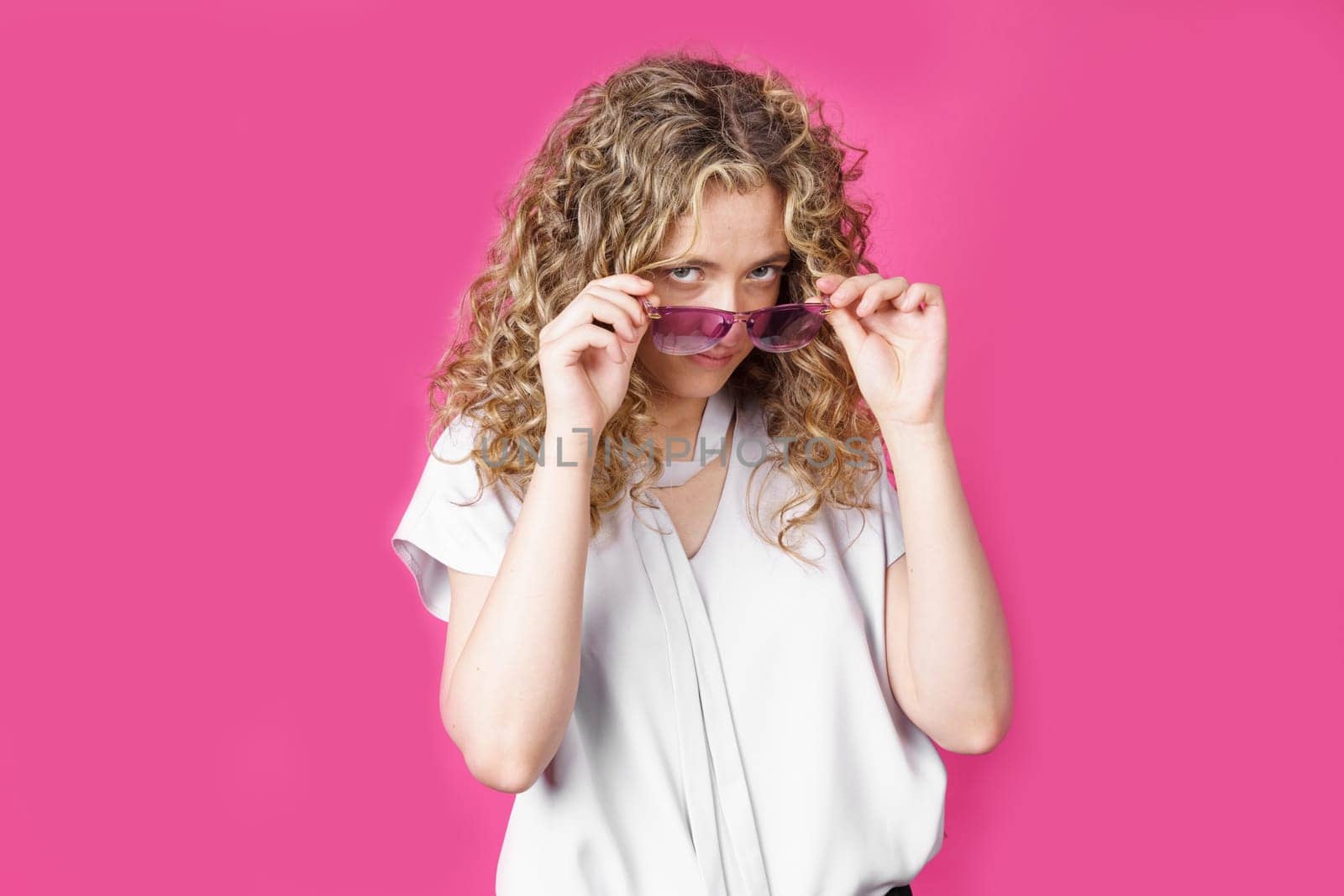 A young woman holds glasses with her hands and looks from under them. Isolated pink background