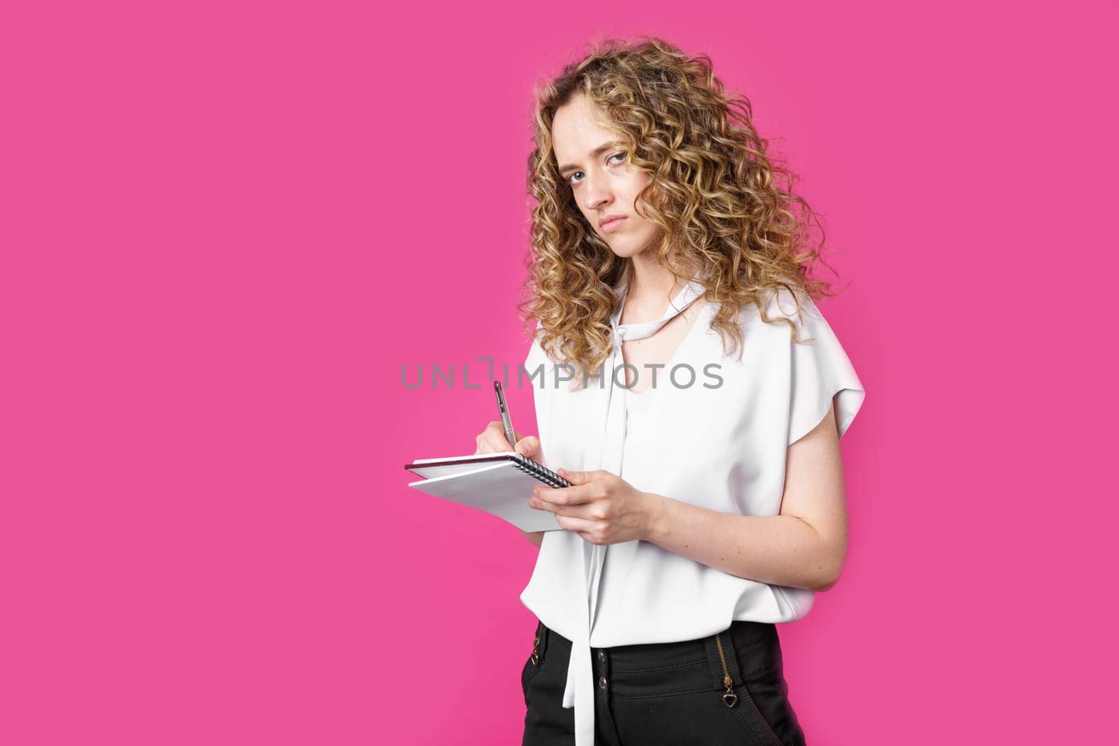 Charming woman makes entries in the diary and looks into the camera. Isolated on a pink background.