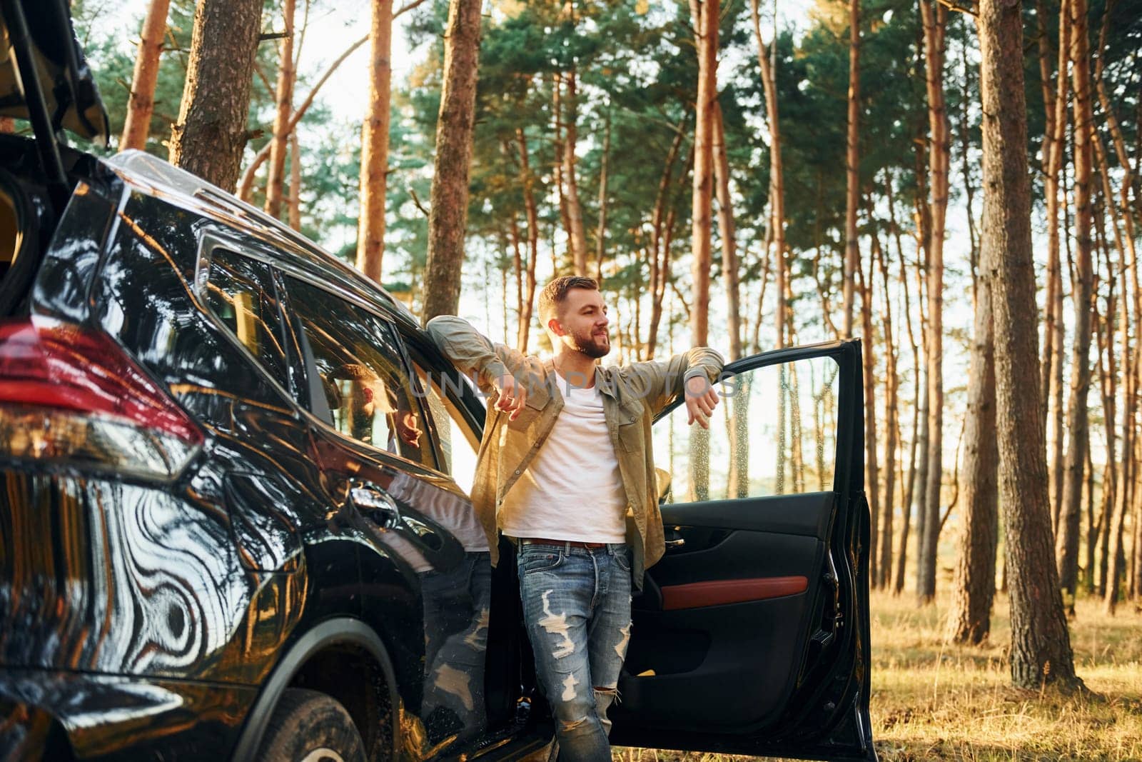 With his car. Man in jeans is outdoors in the forest with his black colored automobile.