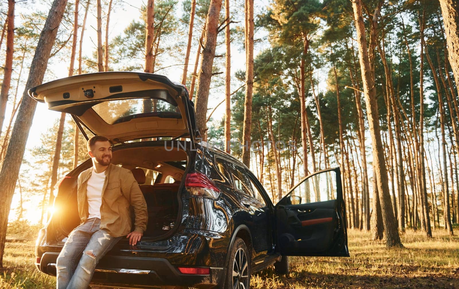 With his car. Man in jeans is outdoors in the forest with his black colored automobile.