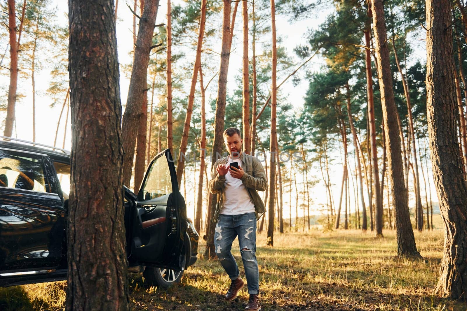 Near the car. Man in jeans is outdoors in the forest with his black colored automobile.