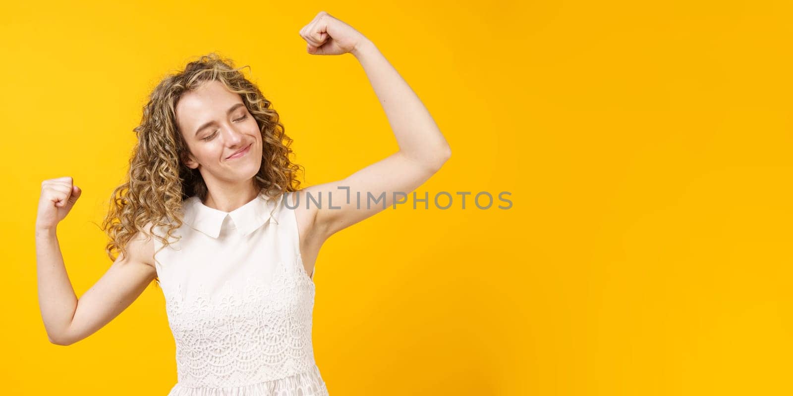 A strong woman with curly hair, a toothy smile, raises her arms and shows her biceps. Models on a yellow background. Look at my muscles
