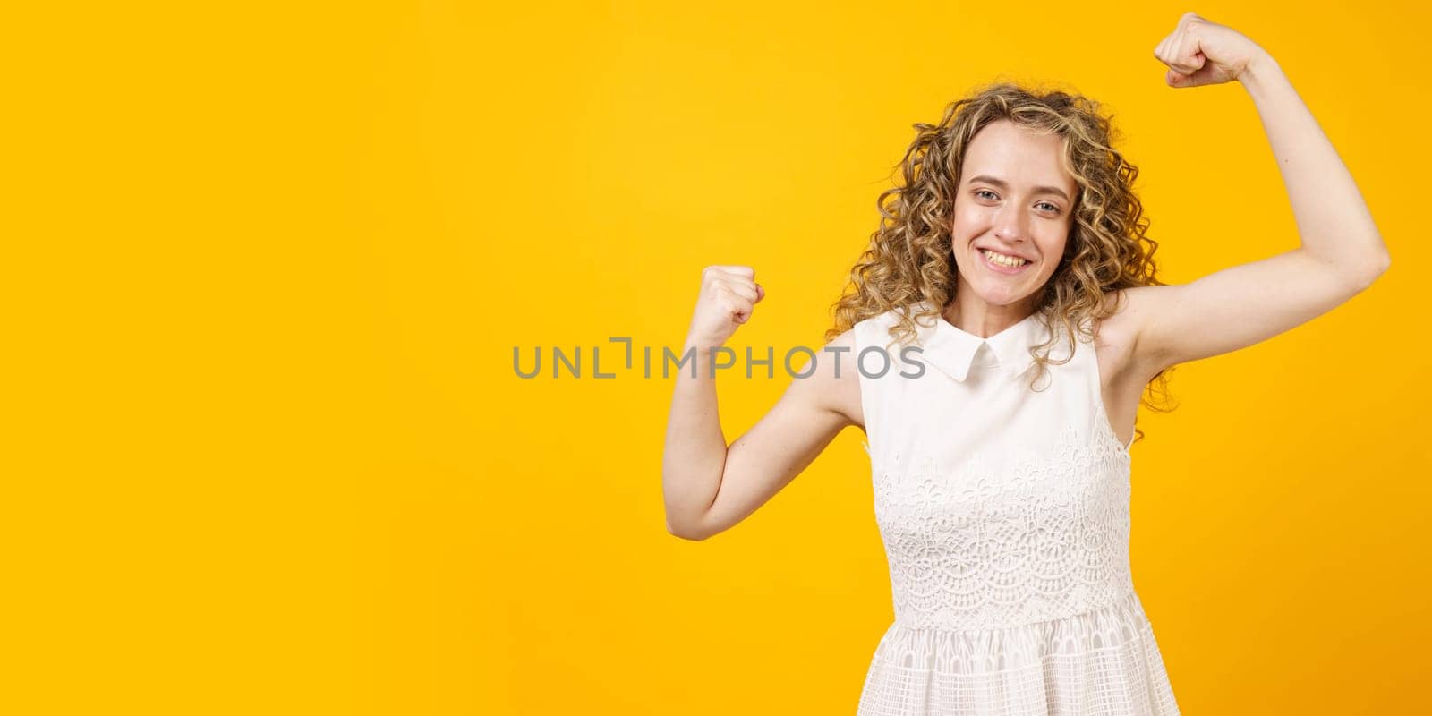 A strong woman with curly hair, a toothy smile, raises her arms and shows her biceps. Models on a yellow background. Look at my muscles