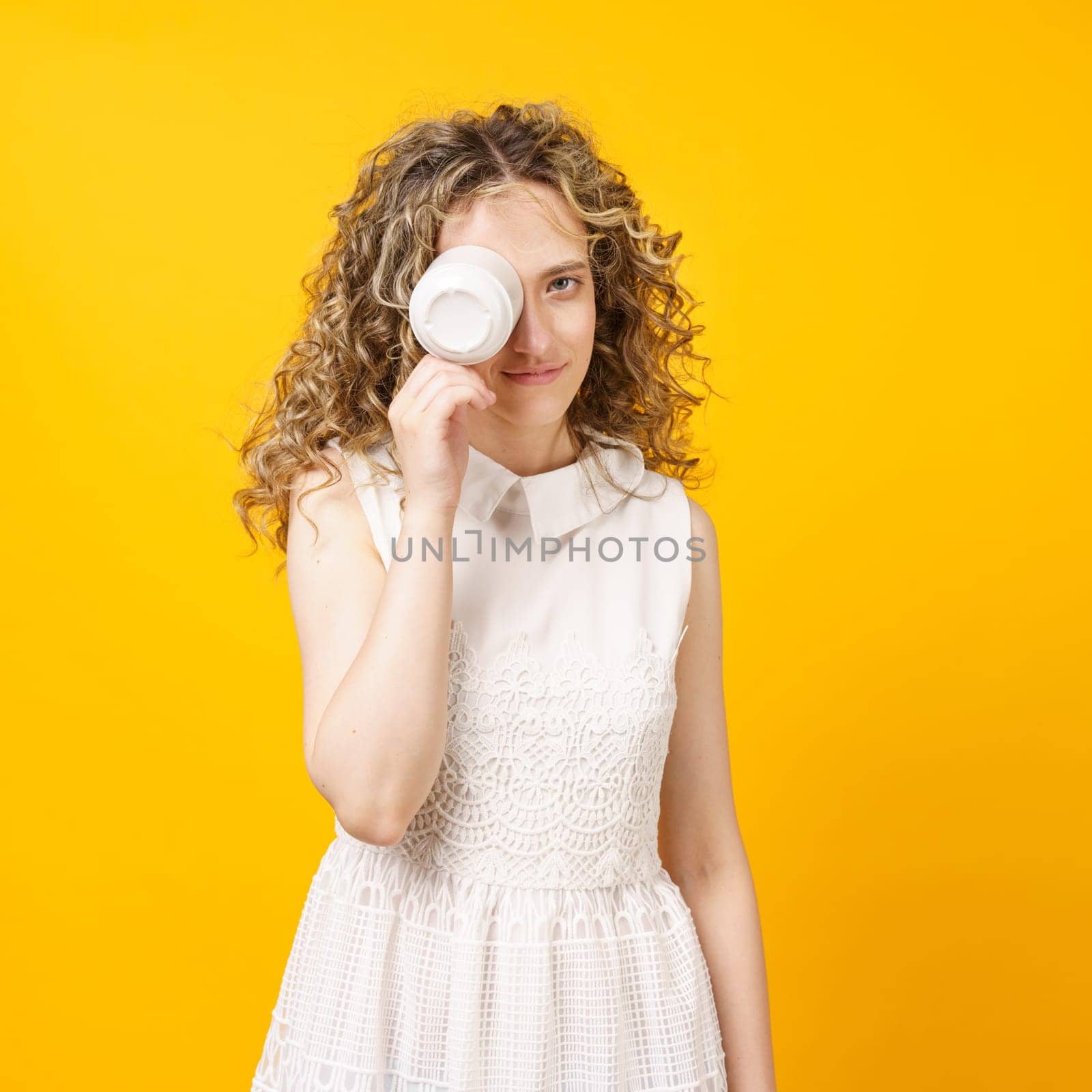 Young woman is covering her face with a cup and smiling. Female portrait. Isolated on yellow background
