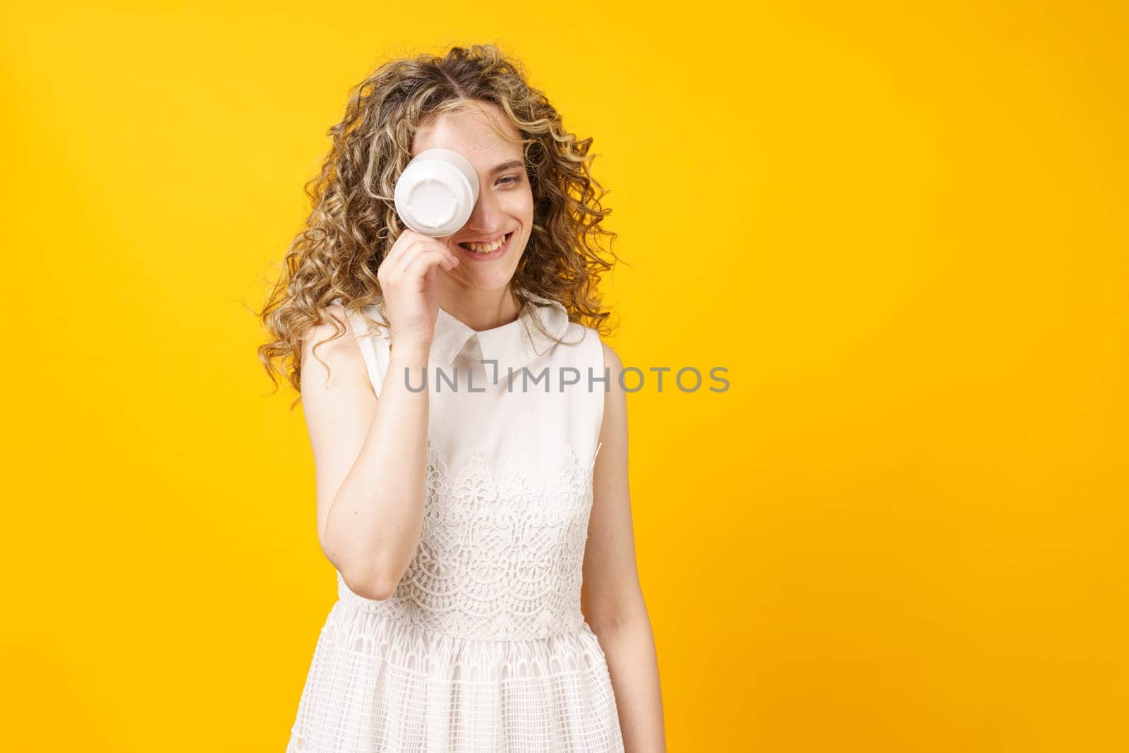 Young woman is covering her face with a cup and smiling. Female portrait. Isolated on yellow background