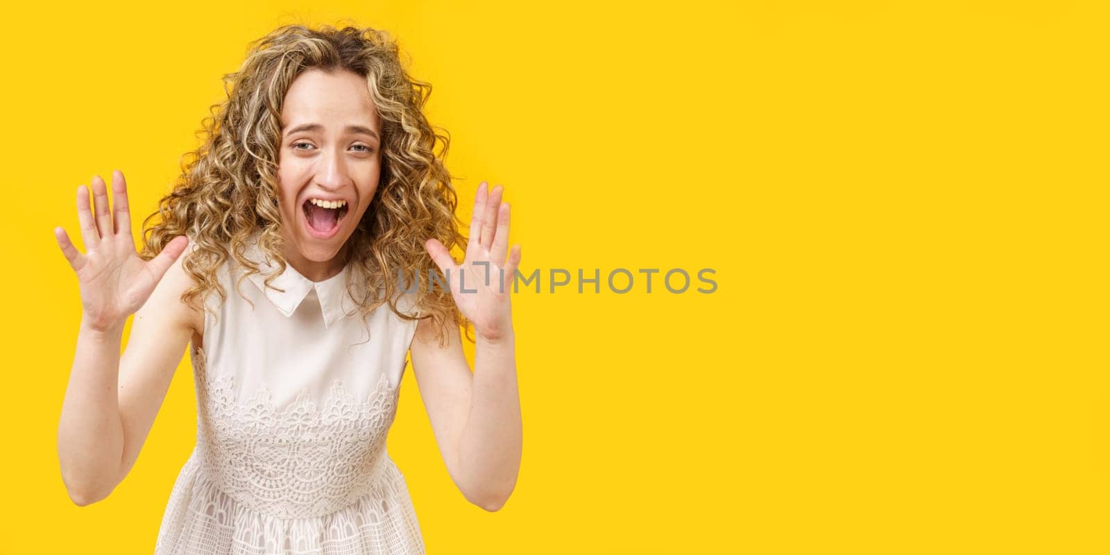A curly-haired woman raises her palms in joy, glad to receive an amazing gift from someone, screams loudly, wearing a striped shirt. Isolated on a yellow background.