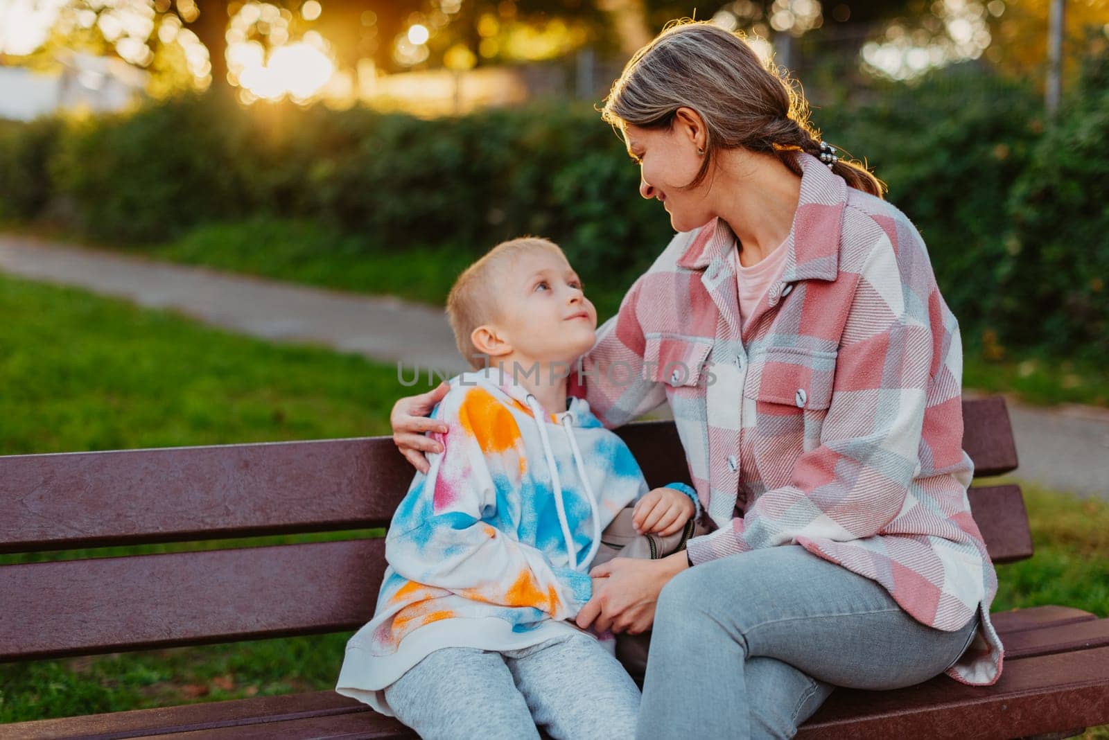 mother and son sit on a park bench in the rays of the setting sun. the concept of a family. Mother's Day. beautiful girl (mother) with a boy (son) in the park in the park are sitting on a bench at sunset.