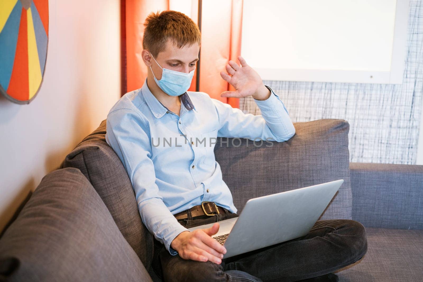 a guy sitting in a protective mask works on the couch via video communication by EkaterinaPereslavtseva